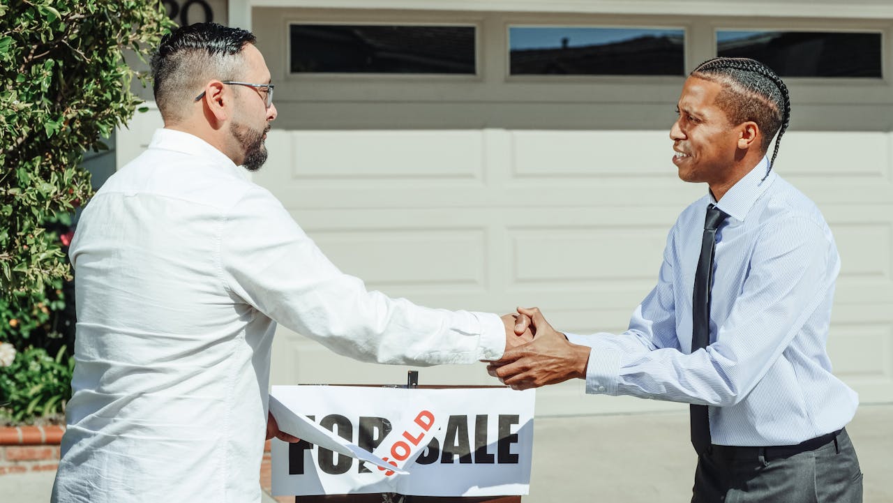 about-01 Two men shaking hands in front of a sold house sign, sealing a real estate transaction.