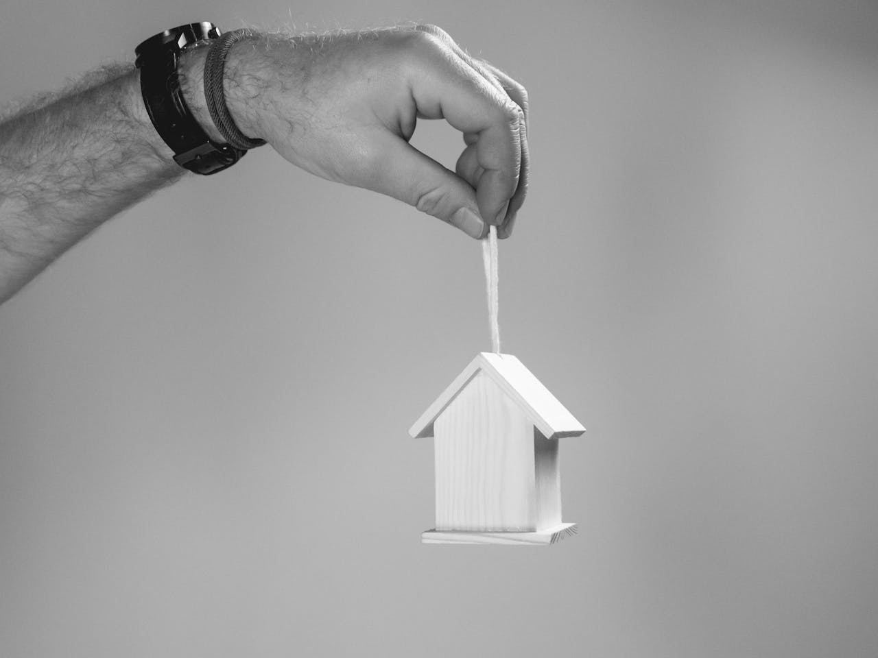 services-02 Black and white photo of a hand holding a miniature wooden house by a string, symbolizing real estate.